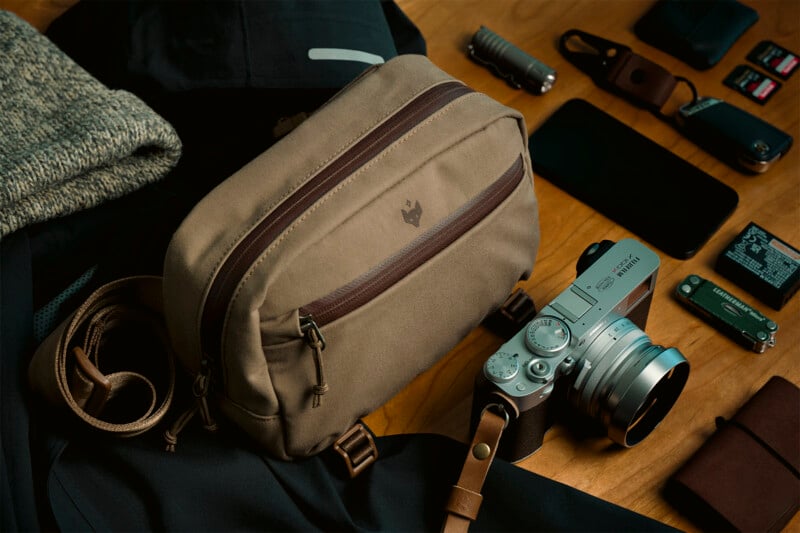 A neatly arranged flat lay of travel essentials on a wooden surface, featuring a tan camera bag, silver digital camera, smartphone, memory cards, flashlight, and clothing items.