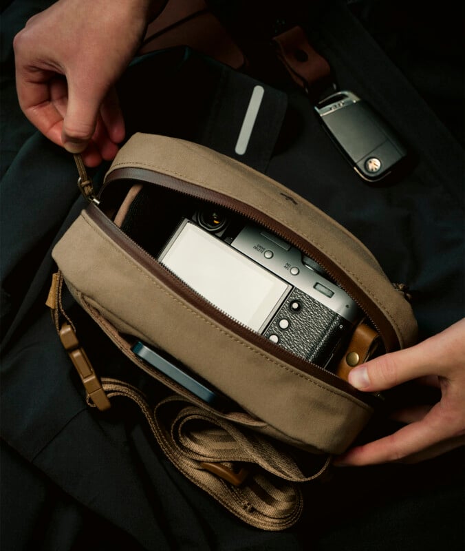 A person places a camera into an open beige canvas bag. The scene includes a set of keys resting nearby on a dark fabric surface. The camera strap is partly visible, indicating casual, on-the-go gear.