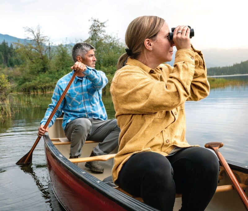 Two people are in a canoe on a calm lake surrounded by trees. The person in the back is paddling, while the person in the front uses binoculars to look at something in the distance. They appear to be enjoying a peaceful day outdoors.