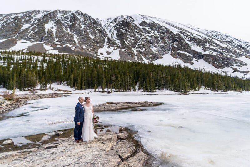 A couple in wedding attire stands on a rocky surface by a partially frozen lake, surrounded by snow-capped mountains and pine trees. The bride holds a bouquet, and they appear to be enjoying the scenic view.