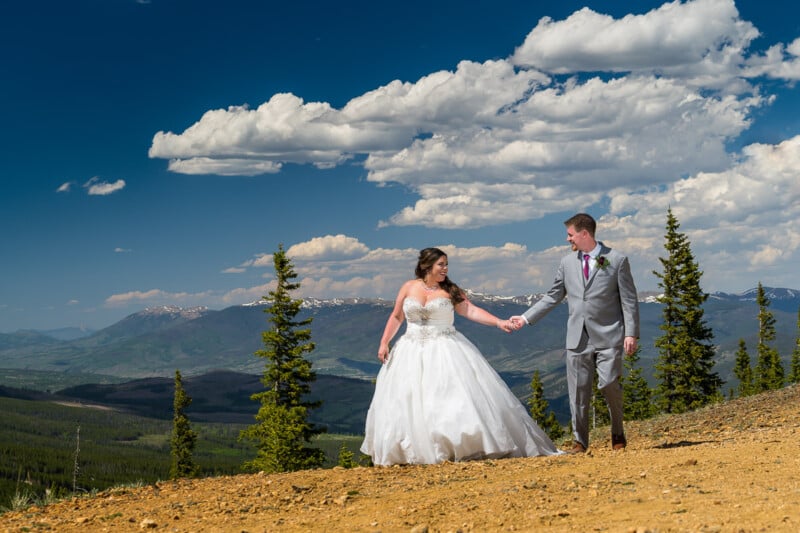 A bride in a white dress and a groom in a gray suit hold hands, walking on a dirt path surrounded by trees, with a backdrop of mountains and a blue sky with clouds.