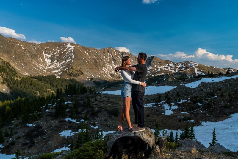 A couple embraces on a rocky outcrop with a scenic mountain landscape in the background. They are surrounded by snow patches and evergreen trees under a clear blue sky.