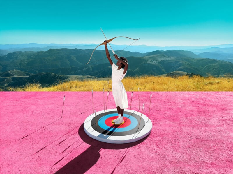 A person in a white outfit stands on a large colorful target painted on a pink surface, aiming a bow and arrow upwards. In the background, there are scenic mountains under a clear blue sky.