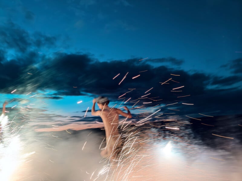 A person runs energetically through colorful, bursting fireworks in the evening sky, creating a dynamic and exhilarating scene against a backdrop of dark clouds.