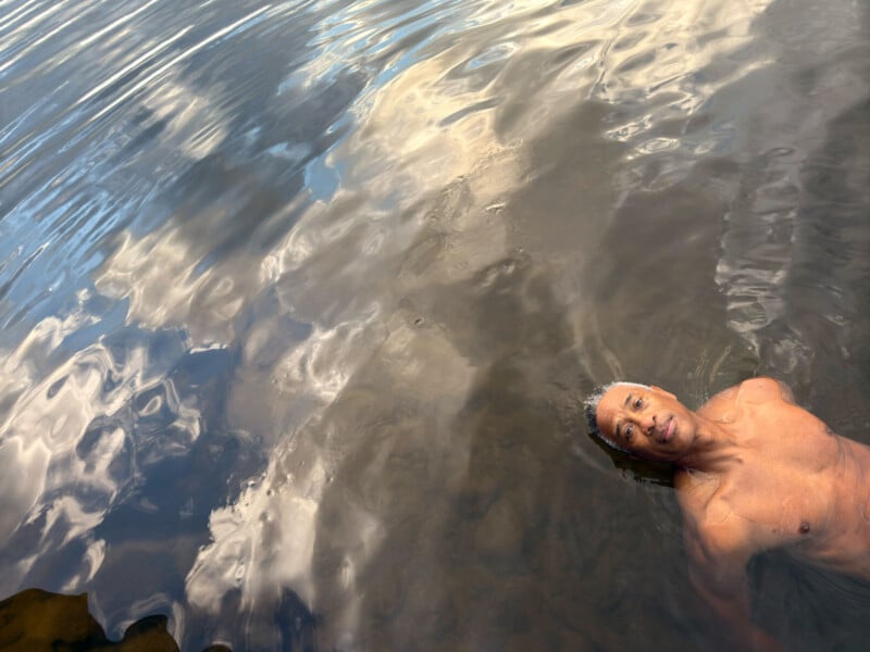 A person with short hair is floating in a calm body of water, looking up. The sky and clouds are reflected on the water's surface, creating a serene and tranquil scene.