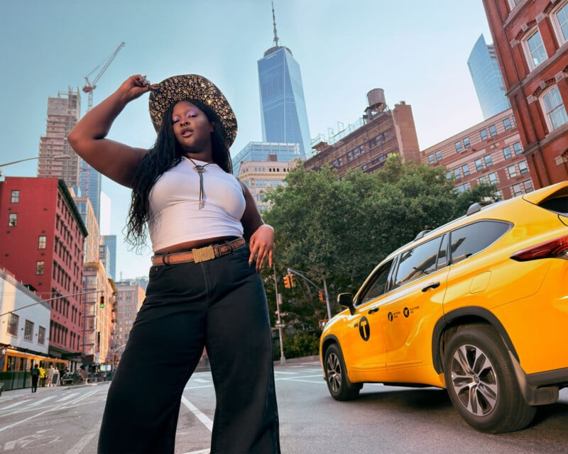 A woman poses confidently in a city street, wearing a hat and white top. A yellow taxi is parked nearby. Tall buildings and a clear sky are in the background, reflecting an urban setting.