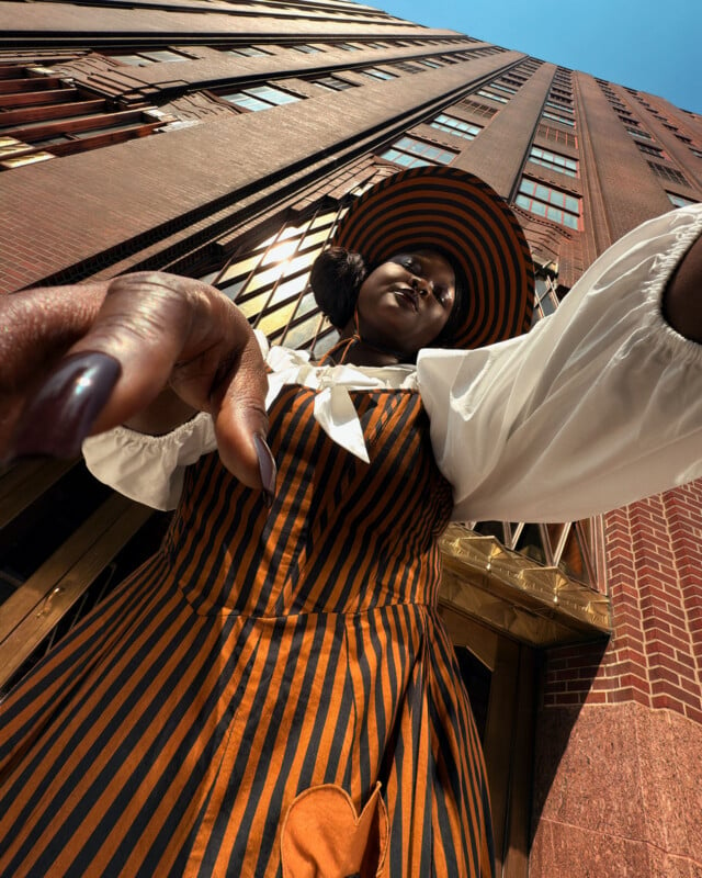 A person in a striped outfit and hat poses confidently with an upward camera angle, emphasizing the tall brick building behind them against a clear blue sky. The outfit features orange and black stripes with a white blouse.