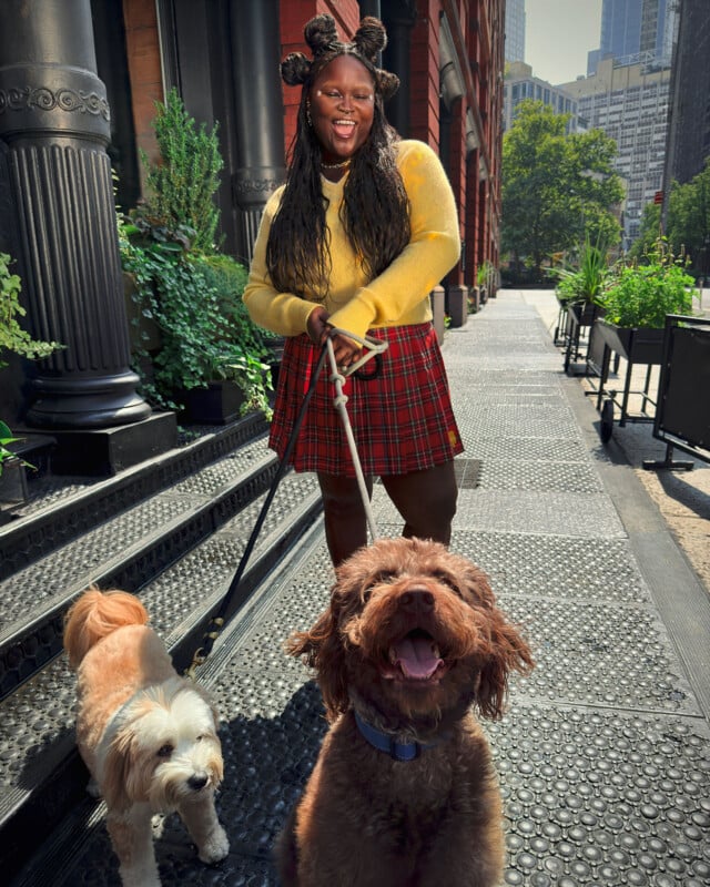 A person with a bright smile stands on a city street, holding the leashes of two happy dogs. They are wearing a yellow sweater and a red plaid skirt. The sidewalk is lined with plants, and tall buildings are visible in the background.