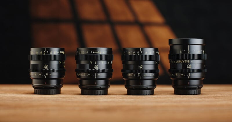 Four black camera lenses with various focal lengths are arranged in a row on a wooden surface, against a warm, softly lit background. Each lens features detailed distance markings in white and yellow.