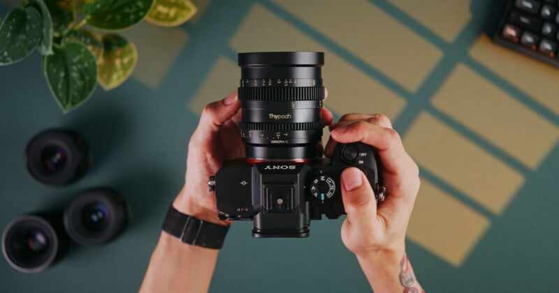 A person holds a Sony camera with a large lens, viewed from above. The table has a few extra lenses, a plant, and part of a keyboard. Shadows create a geometric pattern on the surface.