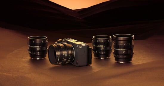 A mirrorless camera with interchangeable lenses is displayed on a sand-like surface, surrounded by four black and gold camera lenses. The background features a gradient of warm brown and orange hues, evoking a sunset in a desert landscape.