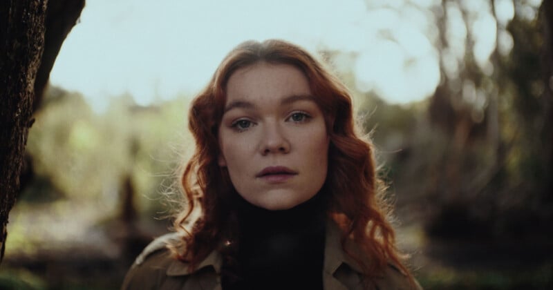 A person with long red hair stands outdoors, looking directly at the camera. They are wearing a dark shirt and a light jacket, with a blurred natural background of trees and sunlight.