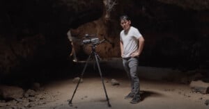 A person stands in a dimly lit cave with a video camera on a tripod. The person is wearing a light gray shirt, dark pants, and sunglasses on their head. The cave's rocky interior is visible in the background.