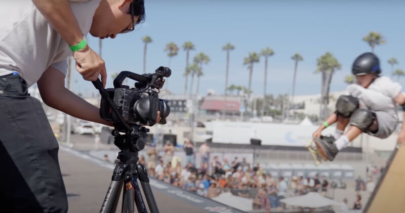 A person operates a camera on a tripod, capturing a skateboarder in mid-air at an outdoor skateboarding event. The background shows a crowd watching and palm trees under a clear sky.