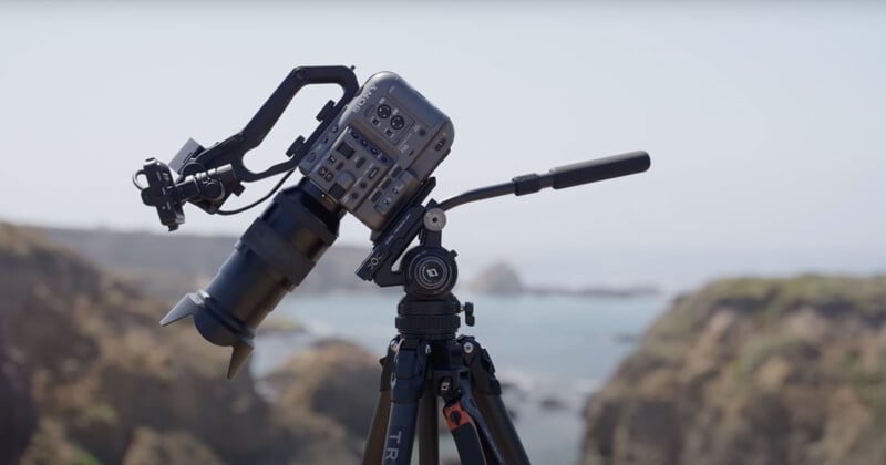 A professional video camera mounted on a tripod is positioned outdoors. The background features a blurred view of the ocean and rocky cliffs under a clear sky.