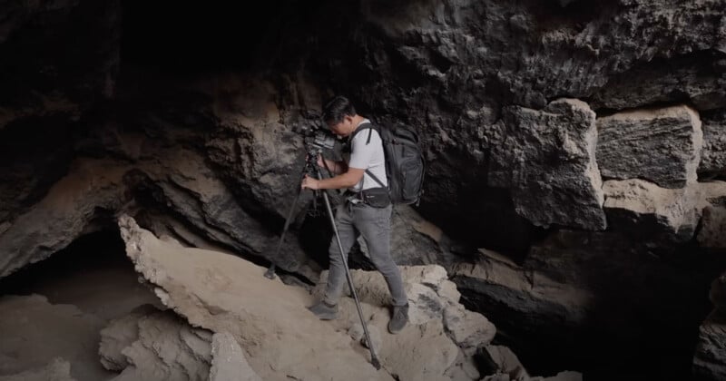 A man with a backpack and tripod carefully walks on uneven rocks inside a dark cave. The rugged cave walls and rocky terrain create a sense of adventure and exploration.