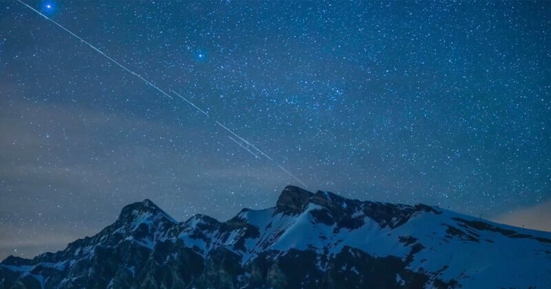 A stunning night sky filled with countless stars over a mountain range with snow-capped peaks. A bright streak, likely a meteor or satellite trail, crosses diagonally, adding a dynamic element to the serene and starry scene.