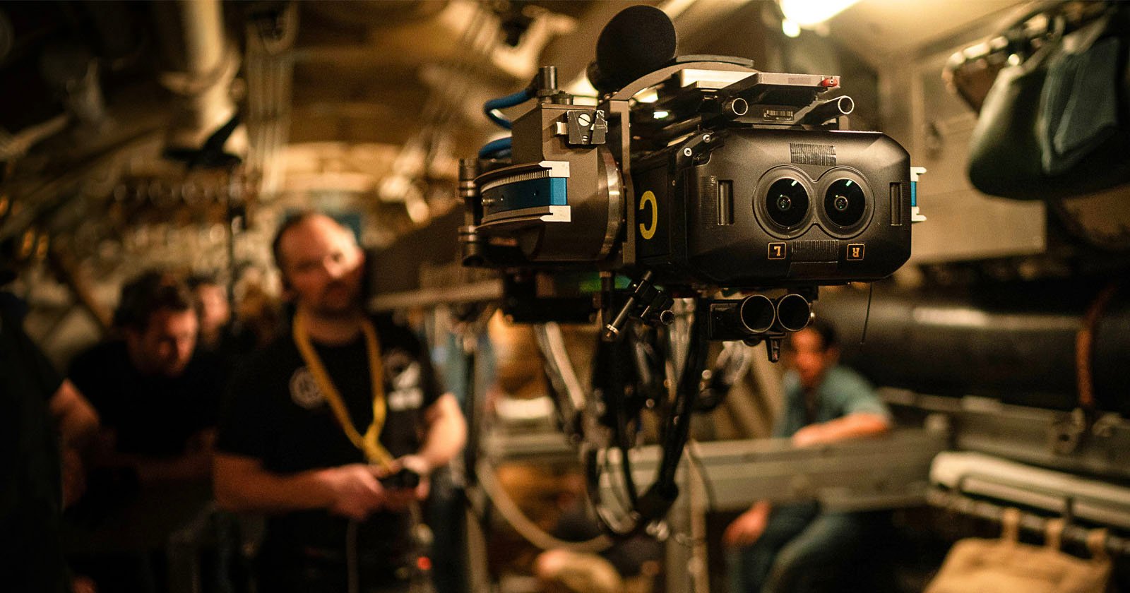 A professional camera rig is set up in a dimly lit industrial or mechanical environment. Several people are in the background, blurred and out of focus, suggesting the setting is a film shoot or a documentary setting.