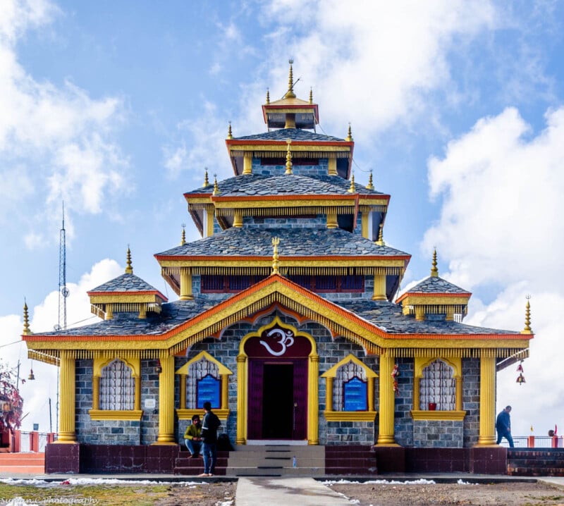 A multi-tiered, traditional temple with colorful details stands under a blue sky. Two people are near the entrance, which features a large Om symbol. The temple is surrounded by a few clouds and has golden spires on its roof.