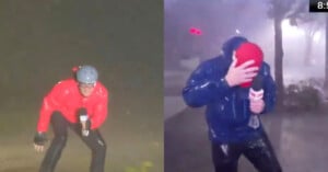 Two people in rain gear report live during a heavy rainstorm. The person on the left crouches with a microphone, wearing a helmet and red jacket. The person on the right braces against the wind, wearing a red cap and blue jacket.