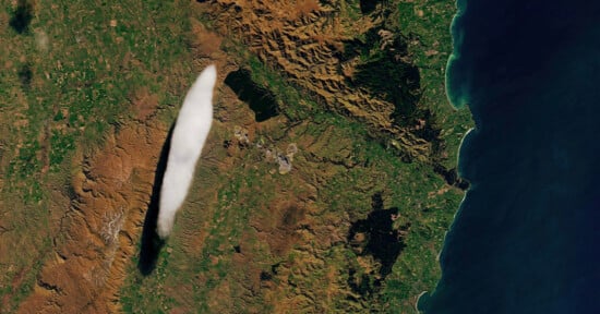 Satellite image showing a long, narrow cloud casting a shadow over a mountainous and green landscape near a coastline. The cloud appears white against the varied shades of green and brown terrain.