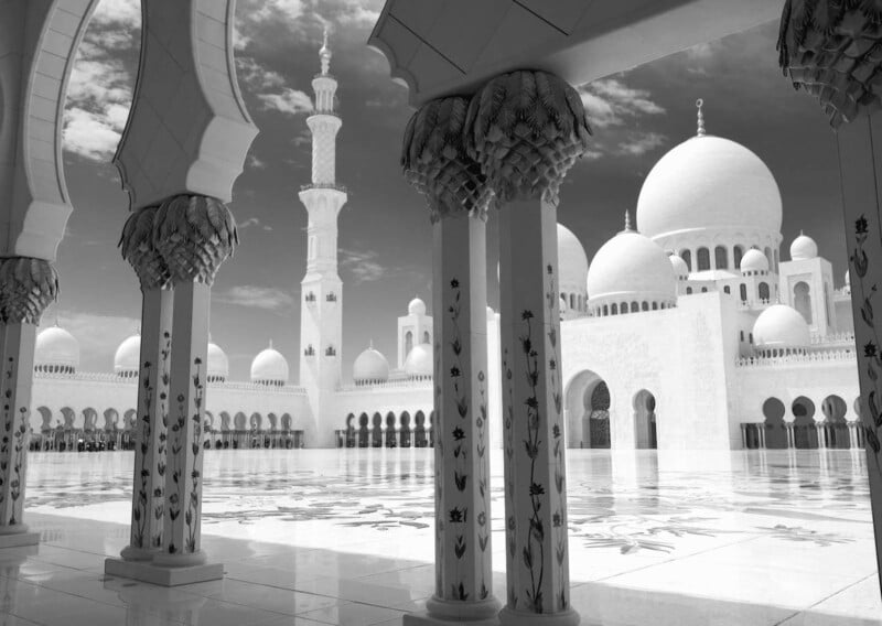 A stunning black and white view of a mosque courtyard, showcasing intricate floral patterns on columns. The domes and minarets stand tall against a partly cloudy sky, reflecting a serene and majestic ambiance.