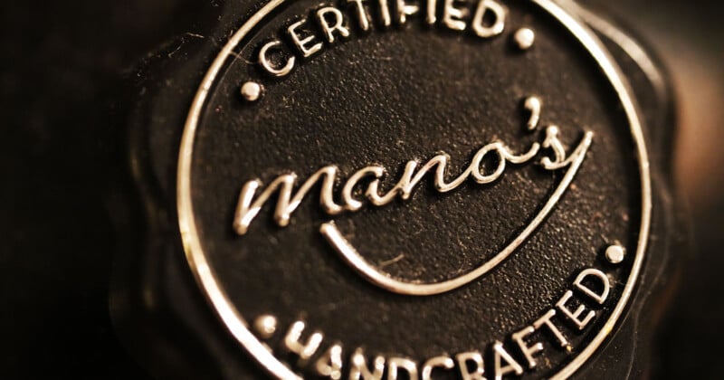 Close-up of a round, dark seal with the words "Certified Handcrafted" encircling the name "mano's" in the center, all embossed in metallic letters. The texture of the seal gives a sense of depth and craftsmanship.