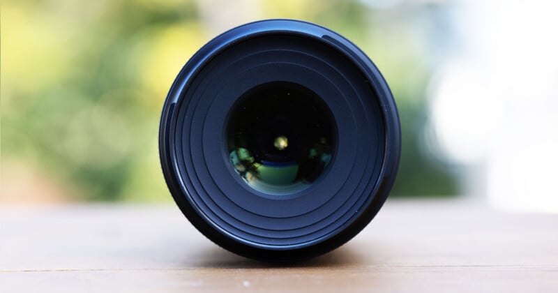 Close-up of a camera lens facing the viewer, placed on a wooden surface. The background is softly blurred with shades of green and white, suggesting an outdoor setting.