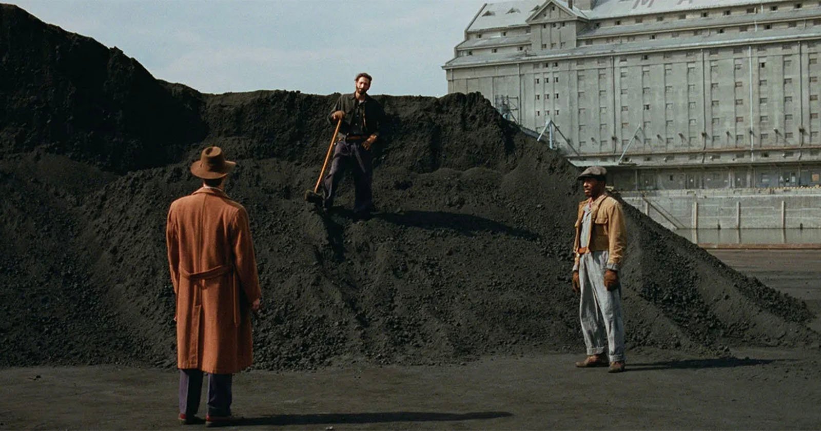 Three men stand on and around a large coal pile. One man stands atop the pile with a shovel, while the other two face him at the base. A large, ornate building is visible in the background. The scene has a vintage, cinematic feel.