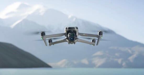 A drone hovers in the foreground against a backdrop of snow-capped mountains and a calm lake. The sky is clear and blue.