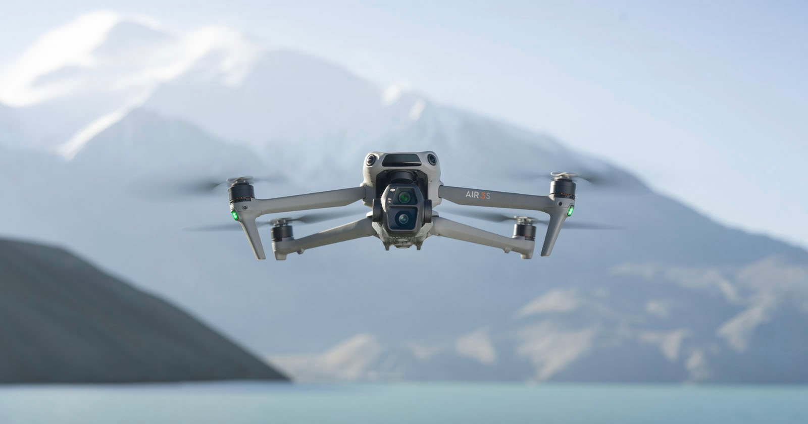 A drone hovers in the foreground against a backdrop of snow-capped mountains and a calm lake. The sky is clear and blue.