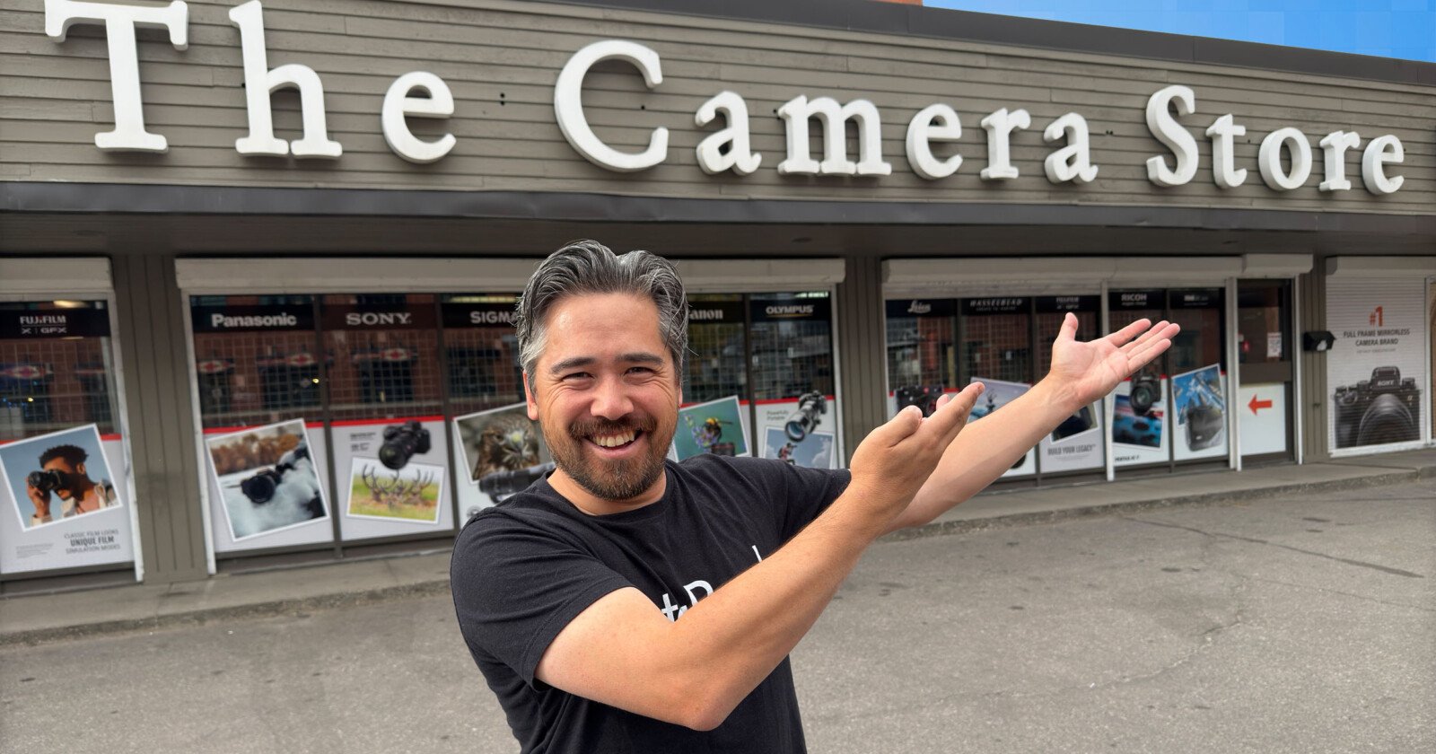 A person with a beard and gray hair, wearing a black t-shirt, smiles while gesturing with both arms in front of "The Camera Store" building, which has window displays of various camera brands and products.