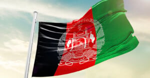 The flag of Afghanistan waves against a clear sky. It features vertical black, red, and green stripes with a white emblem in the center depicting a mosque with flags, sheaves of wheat, and Arabic script.