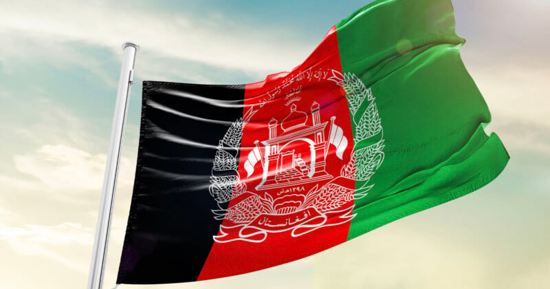 The flag of Afghanistan waves against a clear sky. It features vertical black, red, and green stripes with a white emblem in the center depicting a mosque with flags, sheaves of wheat, and Arabic script.