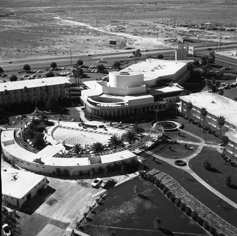 An aerial black-and-white image of a large resort complex surrounded by a desert landscape. The hotel's circular and rectangular structures are visible, along with a pool area and palm trees. The scene includes parking lots and nearby roads.