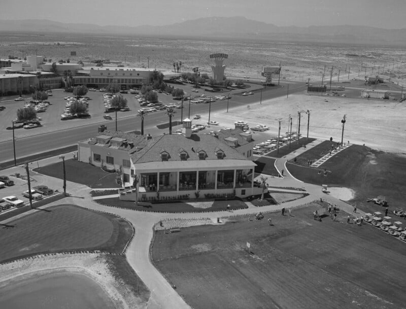 Aerial view of a mid-20th century hotel and golf course in a desert setting, featuring a large building with a terrace, neatly maintained lawns, and several parked cars. The background shows a road, sparse buildings, and distant mountains.