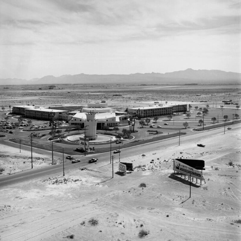 Aerial view of a mid-20th century desert motel complex with a circular central building and surrounding wings. Sparse traffic on nearby roads, a billboard, and distant mountains under a cloudy sky.