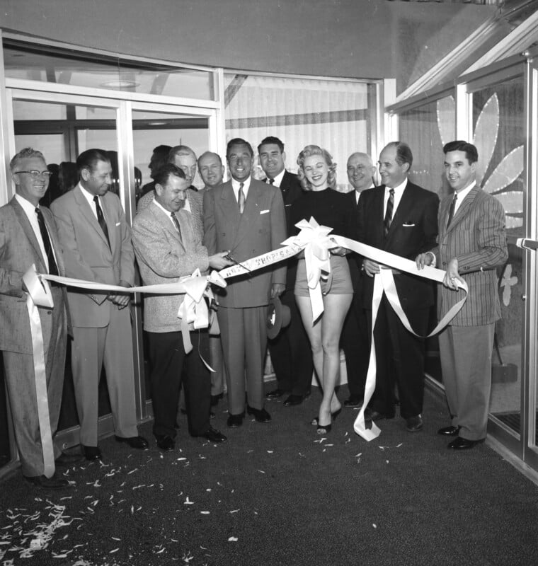 A group of men in suits and a woman in a dress participate in a ribbon-cutting ceremony. The woman is standing in the center, holding scissors to cut the ribbon. Confetti is scattered on the floor, and a glass door is in the background.