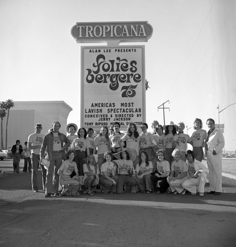 A group of performers pose in front of a large billboard for "Folies Bergere" at the Tropicana. They are wearing matching shirts and standing on a street with palm trees and power lines visible.