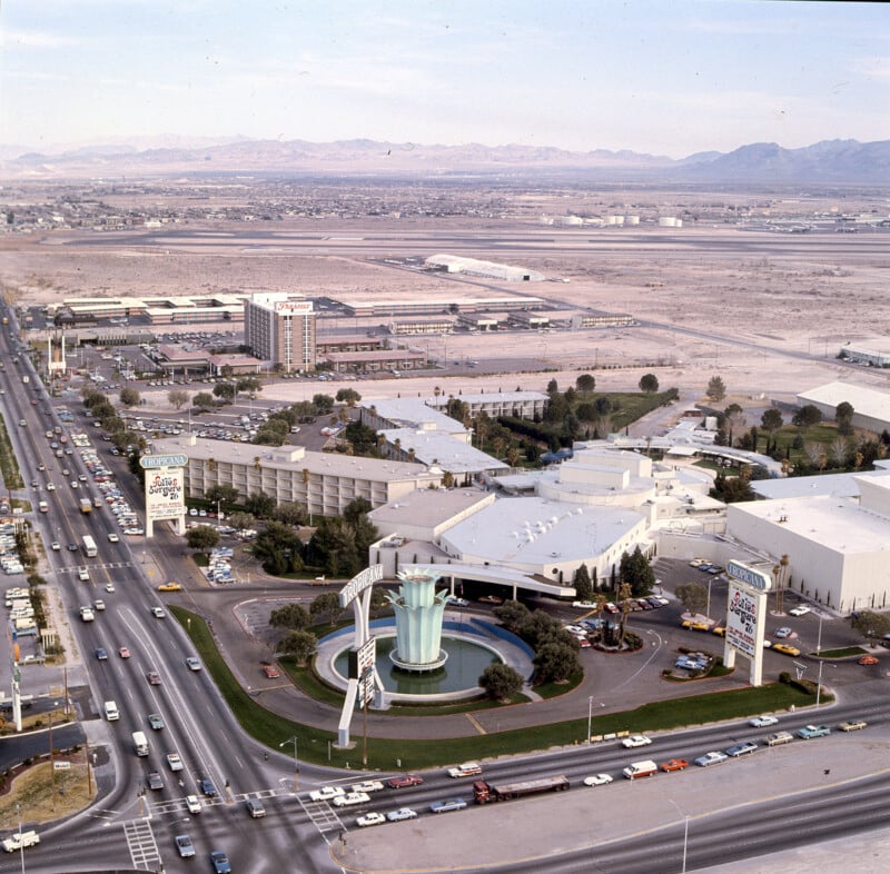 Aerial view of a large building complex surrounded by desert landscape. The complex features a circular fountain and significant signage. Multiple cars travel on the roads, and mountains are visible in the background.