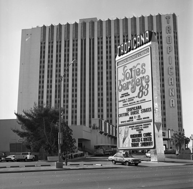 A black-and-white photo of the Tropicana Hotel and Casino in Las Vegas. A large sign in front advertises "Follies Bergere '83" with additional show details. The building is multi-storied with a series of vertical lines on its facade.