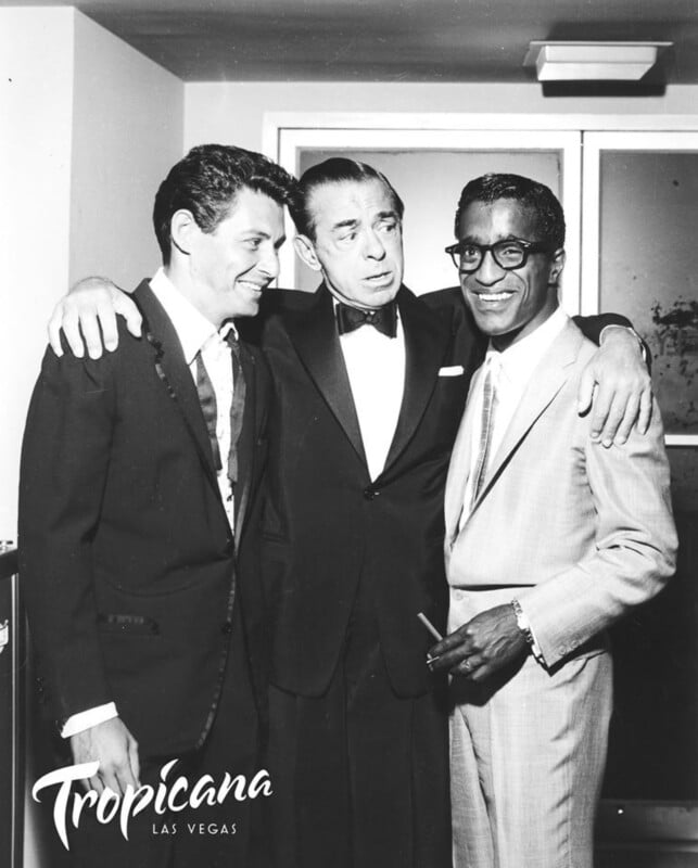 Three men in suits stand together, smiling and engaged in conversation in front of a doorway. One is holding a cigarette. The image is in black and white, with "Tropicana Las Vegas" written at the bottom left corner.