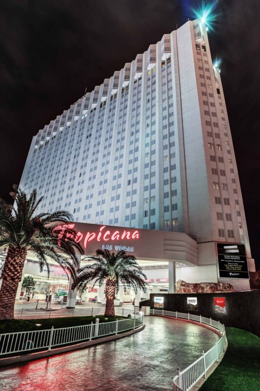 Night view of the Tropicana Las Vegas hotel, a tall building with bright lights illuminating its facade. The entrance is surrounded by palm trees and a paved walkway, with the red and white Tropicana sign prominently displayed.