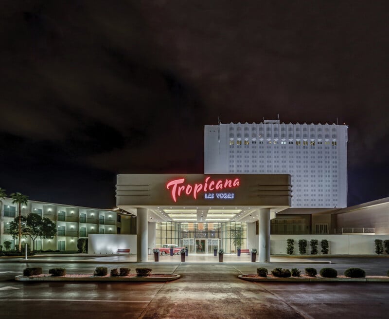 Night view of the Tropicana Las Vegas hotel entrance with bright red neon signage against a dark sky. A multi-story building is in the background, and the foreground is lit by entrance lights.