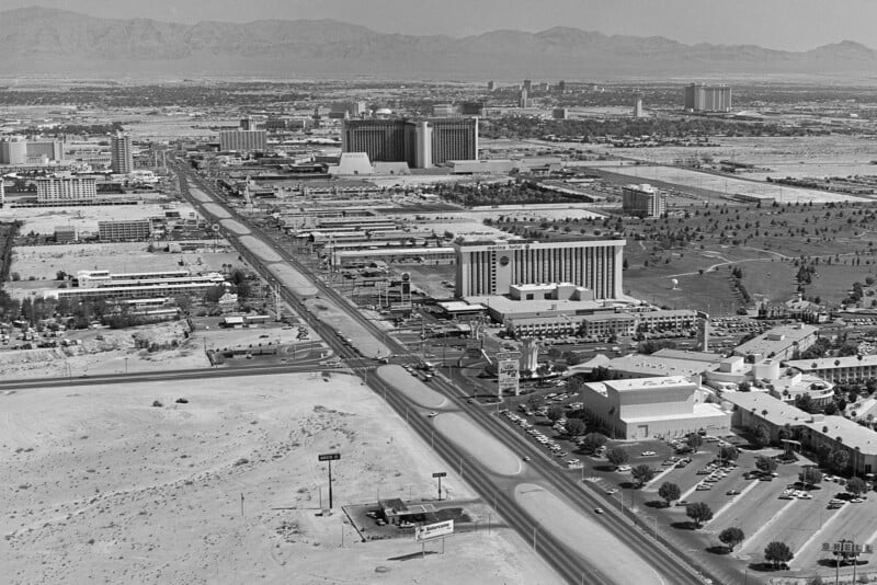 Aerial view of a cityscape with desert surroundings. Wide roads run through the area, flanked by large buildings, including hotels and casinos. Mountains are visible in the distant background, under a clear sky.