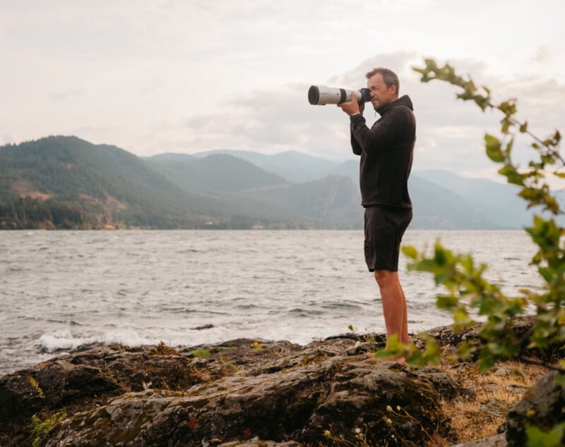 A person stands on rocky terrain by a large body of water, holding a camera with a long lens, aimed at a distant point. They are wearing a black hoodie and shorts. Hills and a cloudy sky form the backdrop. Green foliage is visible in the foreground.