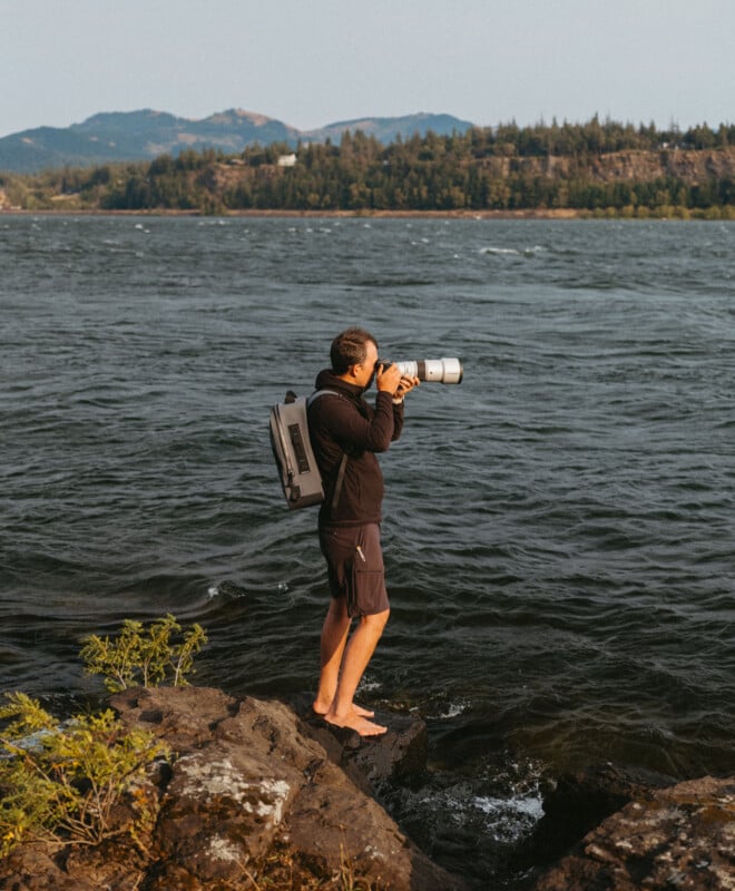 A person stands barefoot on rocks by a lake, wearing a backpack, and photographing with a large zoom lens. The background features forested hills and a partly cloudy sky.