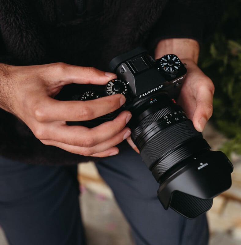 A person adjusts the settings on a Fujifilm camera. The hands are turning a dial on the camera, which features a large lens and lens hood. The background is blurred, highlighting the focus on the camera and hands.