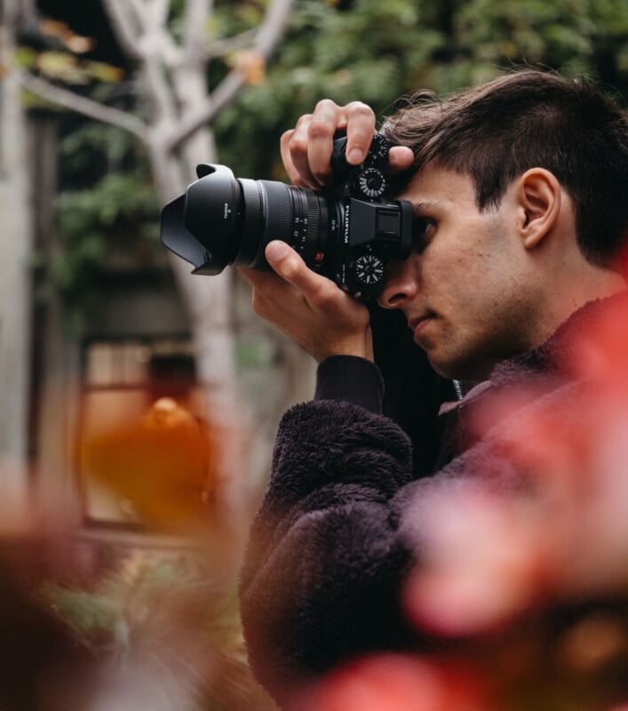 A person wearing a dark jacket is taking a photo with a camera, surrounded by blurred greenery and red foliage. The background is slightly out of focus, highlighting the photographer's concentration and the natural setting.