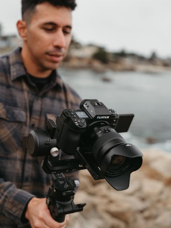 A person holding a Fujifilm camera mounted on a gimbal, standing by a rocky coastline. The background is blurred, highlighting the focus on the camera equipment.